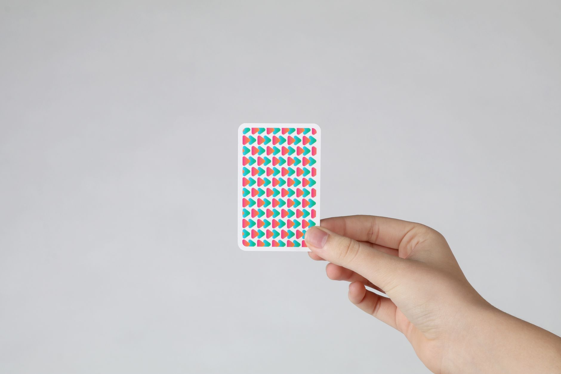 Une main tient une carte à jouer dont le dos affiche un motif de triangles roses et turquoise, sur fond gris clair uni.